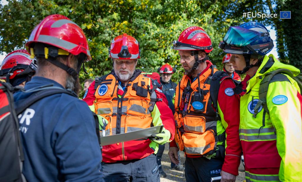 Group of first responders wearing helmets and high-visibility rescue gear gather outdoors, examining a map or planning document. They appear to be coordinating an emergency or training operation in a wooded area.