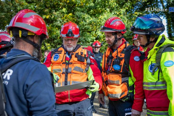 Group of first responders wearing helmets and high-visibility rescue gear gather outdoors, examining a map or planning document. They appear to be coordinating an emergency or training operation in a wooded area.