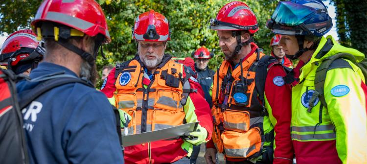 Group of first responders wearing helmets and high-visibility rescue gear gather outdoors, examining a map or planning document. They appear to be coordinating an emergency or training operation in a wooded area.