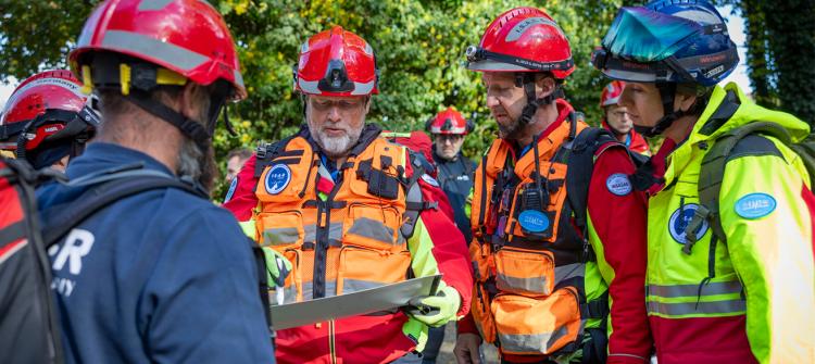 Group of first responders wearing helmets and high-visibility rescue gear gather outdoors, examining a map or planning document. They appear to be coordinating an emergency or training operation in a wooded area.