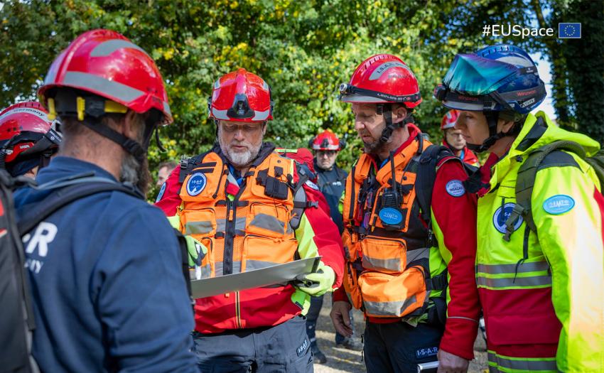 Group of first responders wearing helmets and high-visibility rescue gear gather outdoors, examining a map or planning document. They appear to be coordinating an emergency or training operation in a wooded area.