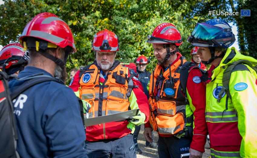 Group of first responders wearing helmets and high-visibility rescue gear gather outdoors, examining a map or planning document. They appear to be coordinating an emergency or training operation in a wooded area.
